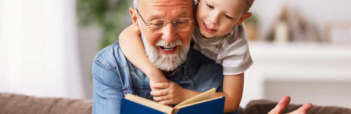 Ein Großvater liest seinem Enkel aus einem Buch vor. Der Enkel umschlingt von hinten seine Schultern.
