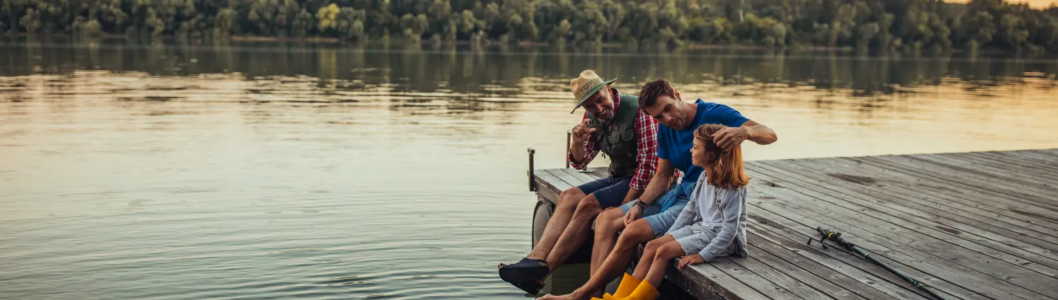 Großvater, Vater und Tochter in Gummistiefeln sitzen an einem See auf dem Steg und lassen die Beine baumeln.