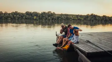 Großvater, Vater und Tochter in Gummistiefeln sitzen an einem See auf dem Steg und lassen die Beine baumeln.