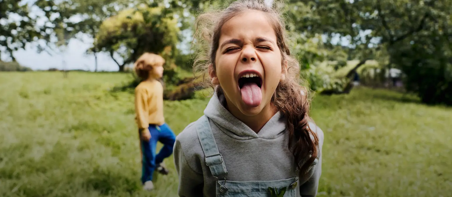 Zwei Kinder toben im Garten: Ein Mädchen stürmt auf die Kamera zu und streckt die Zunge heraus.