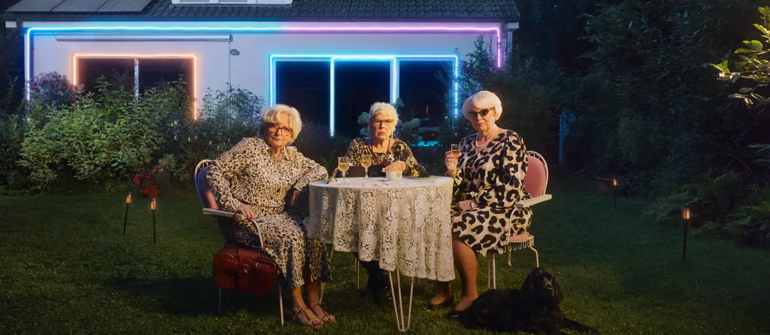 Drei ältere Damen in Kleidern mit Leopardenmuster sitzen an einem Tisch im Garten, spielen Rommé und trinken dabei Wein.