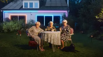 Drei ältere Damen in Kleidern mit Leopardenmuster sitzen an einem Tisch im Garten, spielen Rommé und trinken dabei Wein.