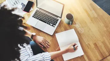 Person von oben, die in ihr Notizbuch schreibt. Daneben eine Tasse, eine Brille und ein Laptop auf dem Tisch.