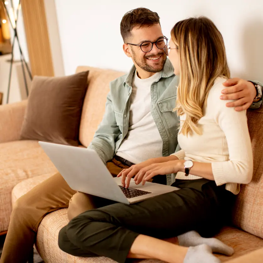 Junges Paar sitzt auf dem Sofa, den Laptop auf den Knien, und schreibt eine Bewerbung.