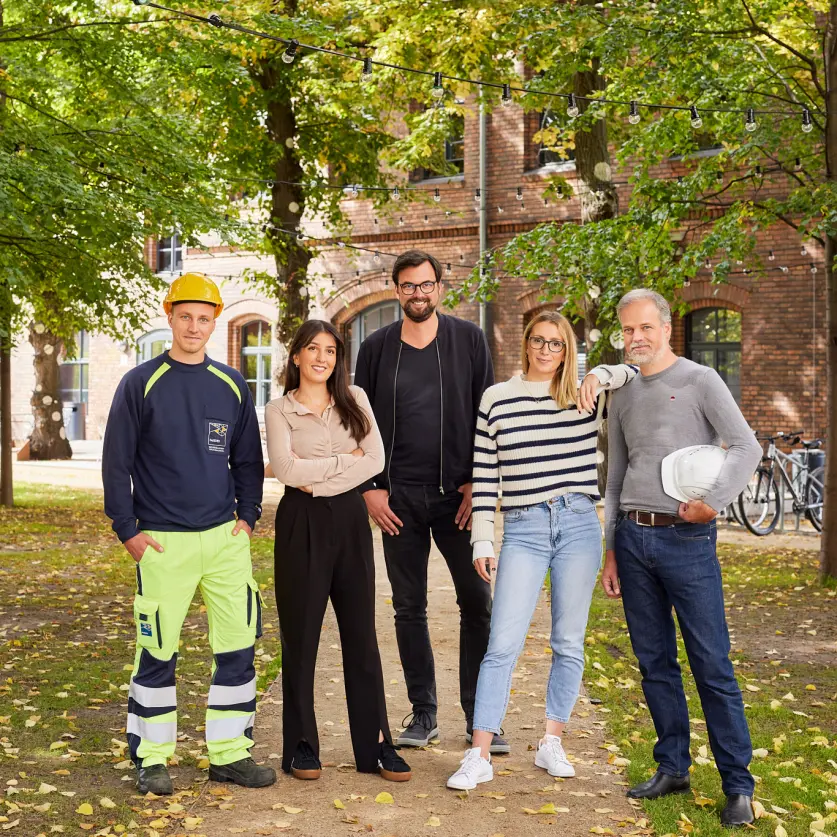 Fünf Mitarbeitende der GASAG-Gruppe in Arbeitskleidung stehen unter Bäumen vor einem Backsteingebäude
