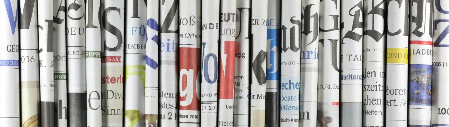In der Mitte gefaltete Zeitungen stehen aufrecht im Regal und bilden ein buntes Mosaik.