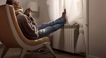 Mann mit Katze auf dem Schoß sitzt im Sessel vor dem Fenster und hat die Füße auf den Heizkörper gelegt.