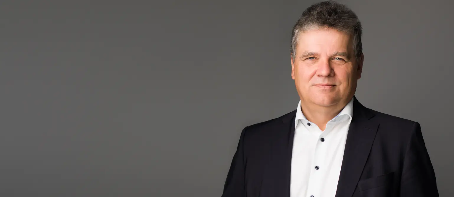 Dr. Jens Horn in dunklem Anzug und weißem Hemd steht vor neutralgrauem Hintergrund und blickt zur Kamera.