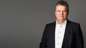 Dr. Jens Horn in dunklem Anzug und weißem Hemd steht vor neutralgrauem Hintergrund und blickt zur Kamera.
