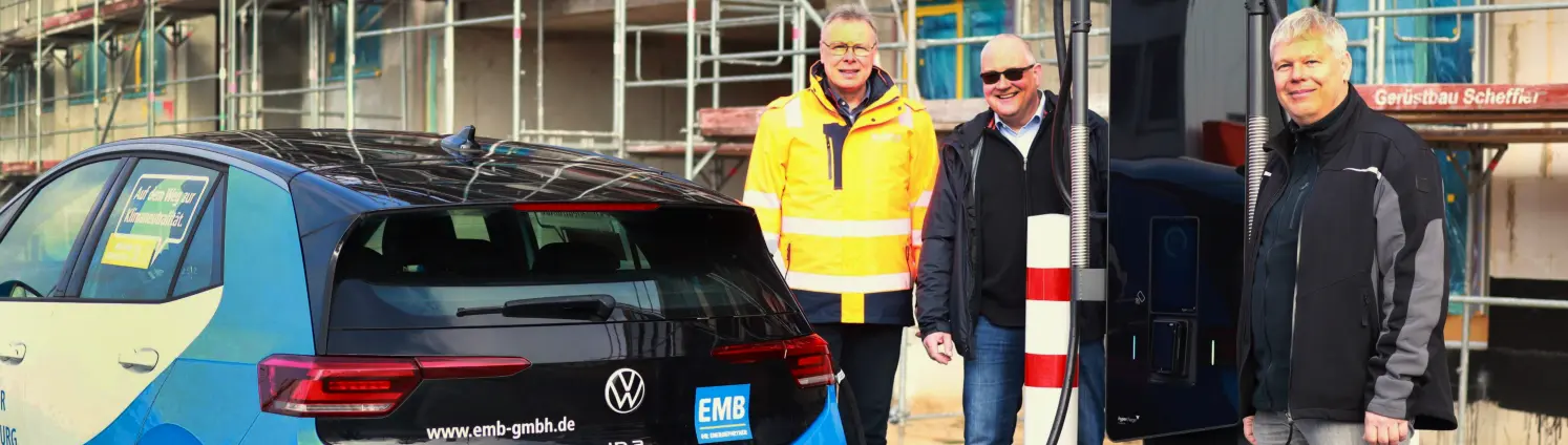 Drei lächelnde Männer stehen neben einer Schnellladestation an der ein blaues Auto gerade auflädt.