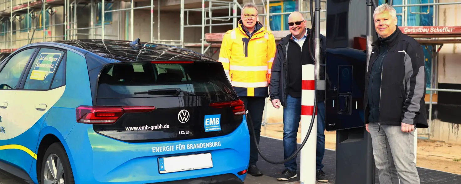 Drei lächelnde Männer stehen neben einer Schnellladestation an der ein blaues Auto gerade auflädt.