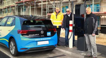 Drei lächelnde Männer stehen neben einer Schnellladestation an der ein blaues Auto gerade auflädt.