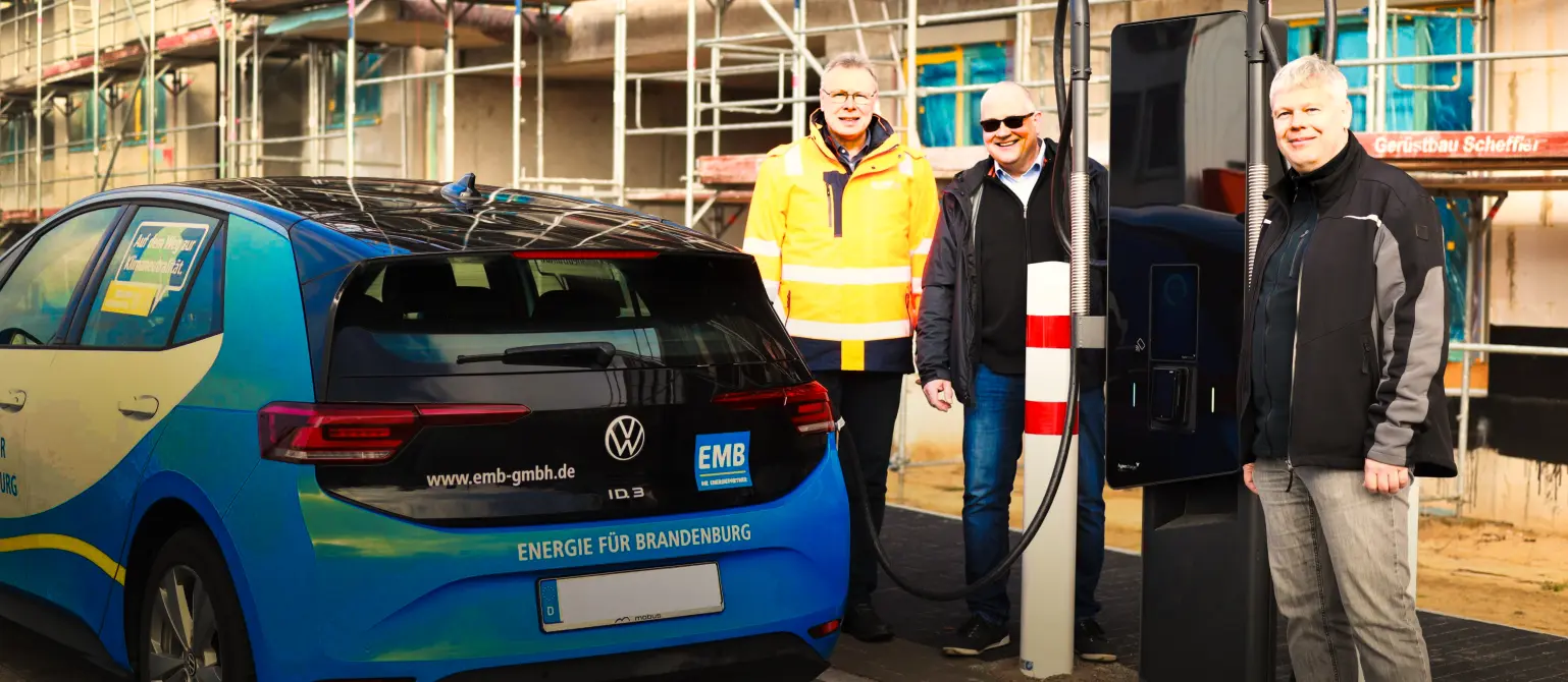 Drei lächelnde Männer stehen neben einer Schnellladestation an der ein blaues Auto gerade auflädt.