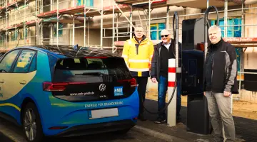 Drei lächelnde Männer stehen neben einer Schnellladestation an der ein blaues Auto gerade auflädt.
