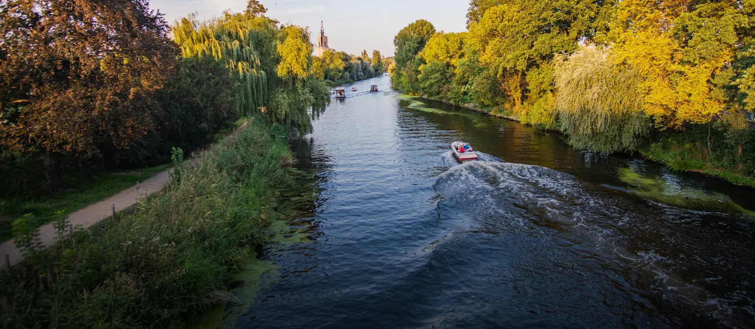 Ein Boot fährt auf einem Fluss, links des Flusses führt ein Fußweg entlang