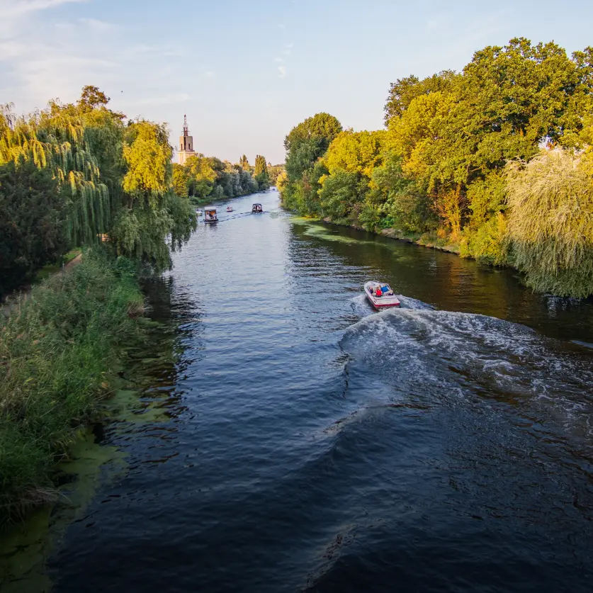 Ein Boot fährt auf einem Fluss, links des Flusses führt ein Fußweg entlang