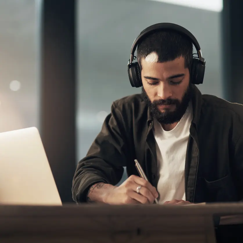 Zahlen und Fakten recherchieren: Ein junger Mann mit Kopfhörer sitzt vor dem Laptop am Schreibtisch und macht sich Notizen.